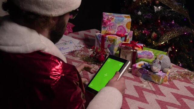 Santa Claus is using the green screen of the tablet for shopping. Tablet with a green screen. Santa Claus sits at a table with a tablet in his hands and flips through web pages.chroma key.