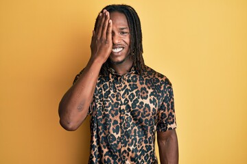African american man with braids wearing leopard animal print shirt covering one eye with hand, confident smile on face and surprise emotion.