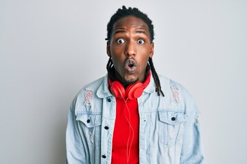 African american man with braids listening to music using headphones scared and amazed with open mouth for surprise, disbelief face