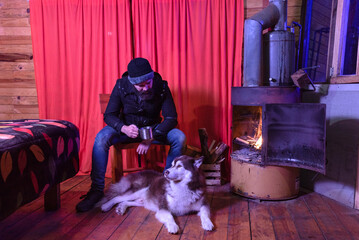 Obraz na płótnie Canvas Bearded lumberjack is inside a log cabin drinking tea from a metal mug sitting next to his husky dog ​​enjoying the warmth provided by an old fireplace made from a rusty barrel with burning firewood