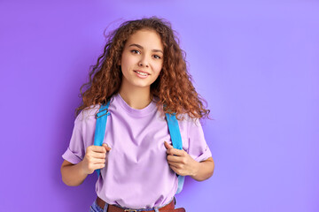 diligent shy caucasian school girl carrying school bag posing at camera, intelligent female teen isolated on purple background