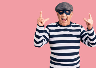 Senior handsome man wearing burglar mask and t-shirt shouting with crazy expression doing rock symbol with hands up. music star. heavy music concept.
