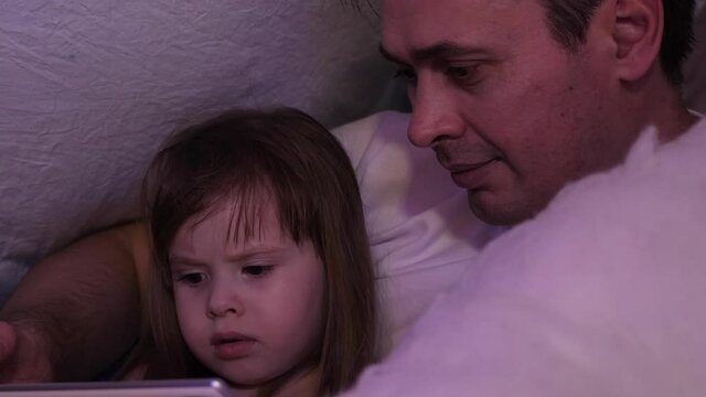 Dad and daughter on Christmas evening play and watch cartoons on a tablet, in a tent with garlands in nursery. Baby and dad are playing in the room. Happy childhood and family concept. New Year