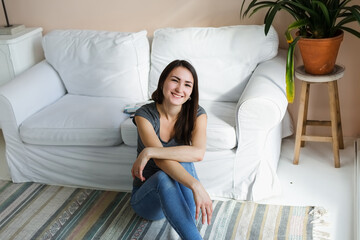 Naklejka premium Girl in jeans sits on the floor, near the sofa, in a bright room.