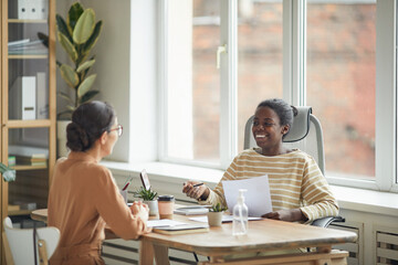 Portrait of smiling African-American woman talking to young woman across table during job interview in office, copy space - obrazy, fototapety, plakaty