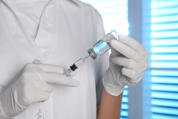 Doctor filling syringe with vaccine from vial indoors, closeup