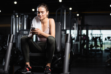 sportswoman sitting on training machine, chatting on smartphone and listening music in wireless earphone