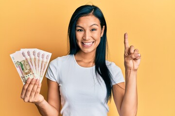 Beautiful hispanic woman holding 100 russian ruble banknotes smiling with an idea or question pointing finger with happy face, number one