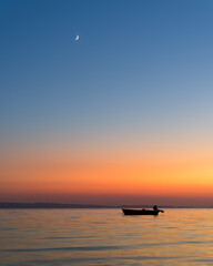Small boat anchored in the calm sea during late burning colorful sunset and a crescent moon visible on the blue sky in Greece , Neos Marmaras 
