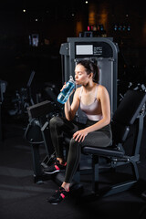 Fototapeta na wymiar young sportswoman sitting on training machine with closed eyes and drinking from sports bottle