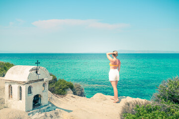A girl wearing white shorts topless standing on a rock looking into the sea distance and dreaming on a sunny day near the small white temple in Greece 