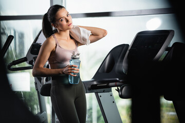 tired sportswoman standing on treadmill, wiping neck with towel and holding sports bottle on...