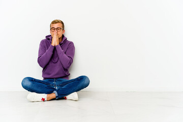 Young caucasian man sitting on the floor holding hands in pray near mouth, feels confident.