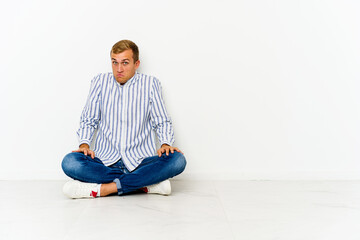 Young caucasian man sitting on the floor shrugs shoulders and open eyes confused.