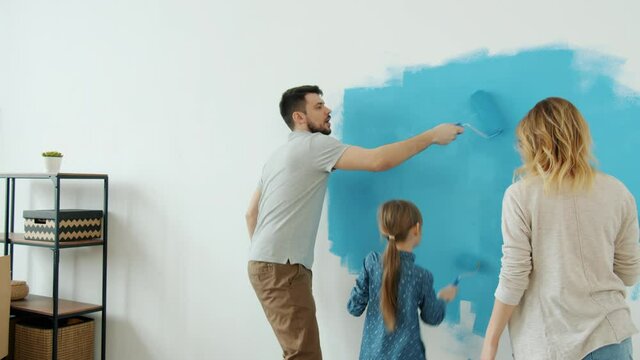 Joyful family with kid remodelling house painting wall and dancing enjoying music together. Happy young people and interior decoration concept.