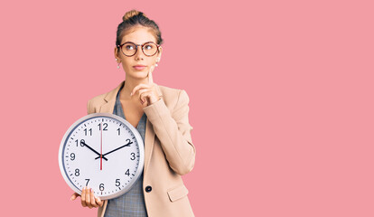 Beautiful caucasian woman with blonde hair wearing business jacket and holding clock serious face...