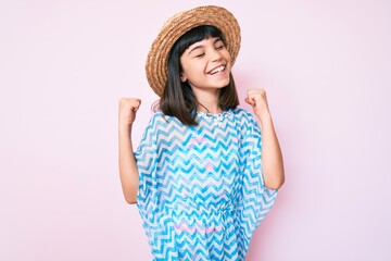 Young little girl with bang wearing summer dress and hat very happy and excited doing winner gesture with arms raised, smiling and screaming for success. celebration concept.