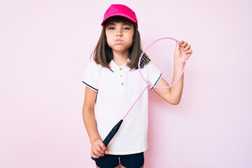 Young little girl with bang holding badminton racket puffing cheeks with funny face. mouth inflated with air, catching air.