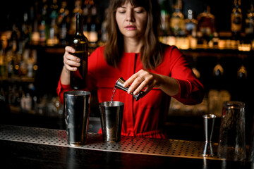 woman bartender pours liquid from jigger into steel shaker cup standing on bar