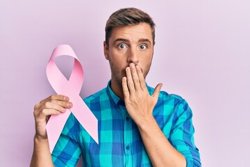 Handsome caucasian man holding pink cancer ribbon covering mouth with hand, shocked and afraid for...