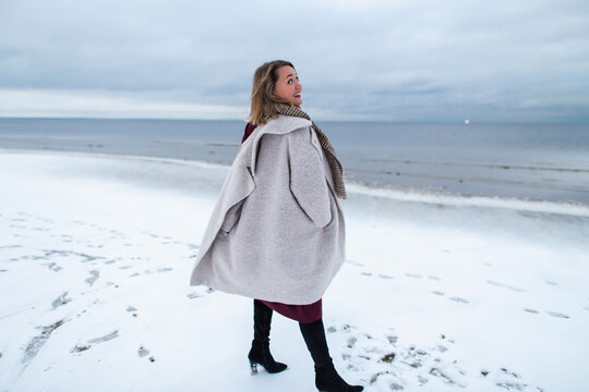Smiling girl in the burgundy dress and coat on the background of the winter sea. Portrait of a woman on sea, snow windy weather, cold atmospheric image.