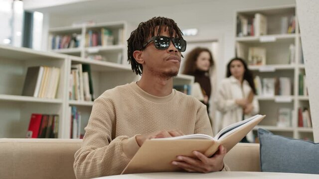 Chest up of young African blind man wearing sunglasses, sitting on couch in public library, reading book with Braille text, moving fingers, blurred female students walking by