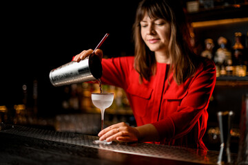 female bartender pours drink from steel glass into frozen glass on bar counter