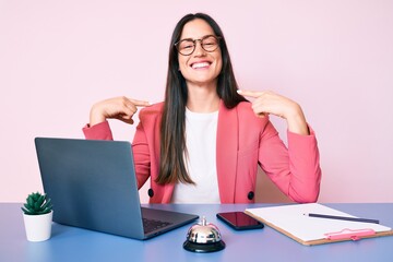 Young caucasian woman sitting at the recepcionist desk working using laptop looking confident with smile on face, pointing oneself with fingers proud and happy.