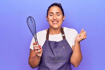 Young beautiful brunette woman wearing apron holding whisk screaming proud, celebrating victory and success very excited with raised arm