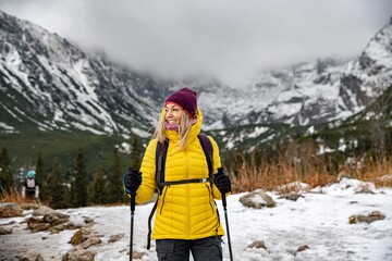 Blonde, smiling traveler woman in down jacket hiking with nordic walking poles on path in mountains.