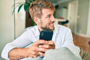 Handsome caucasian man smiling happy sitting on the sofa at home using smartphone