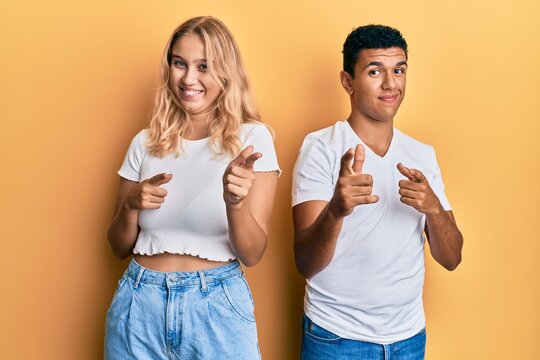 Young interracial couple wearing casual white tshirt pointing fingers to camera with happy and funny face. good energy and vibes.