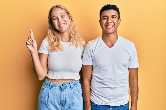 Young interracial couple wearing casual white tshirt showing and pointing up with finger number one while smiling confident and happy.