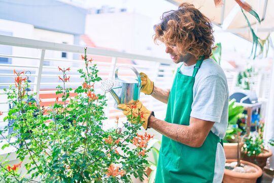 Young hispanic gardener smiling happy caring plants using watering can at terrace