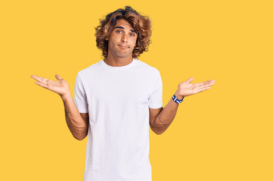 Young hispanic man wearing casual white tshirt clueless and confused expression with arms and hands raised. doubt concept.
