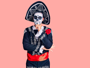 Young man wearing day of the dead costume over background looking confident at the camera with smile with crossed arms and hand raised on chin. thinking positive.