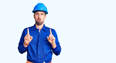 Young handsome man wearing worker uniform and hardhat pointing down looking sad and upset, indicating direction with fingers, unhappy and depressed.