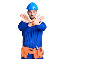 Young handsome man wearing worker uniform and hardhat holding symbolic gun with hand gesture, playing killing shooting weapons, angry face