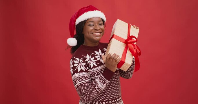 Curious black lady in santa hat shaking gift box, enjoying Christmas holiday, red background