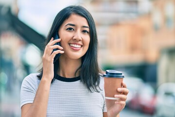 Young hispanic woman talking on the smartphone and drinking take away coffee at the city.