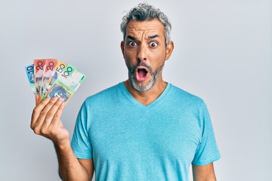 Middle age grey-haired man holding australian dollars scared and amazed with open mouth for surprise, disbelief face