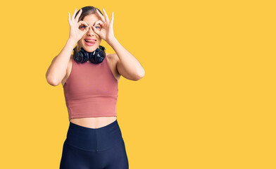 Beautiful caucasian young woman wearing gym clothes and using headphones doing ok gesture like binoculars sticking tongue out, eyes looking through fingers. crazy expression.