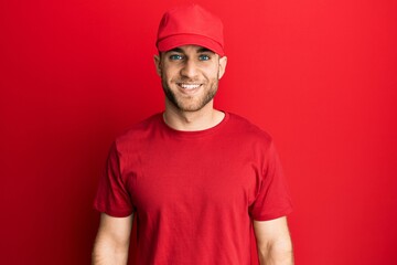 Young caucasian man wearing delivery uniform and cap looking positive and happy standing and smiling with a confident smile showing teeth