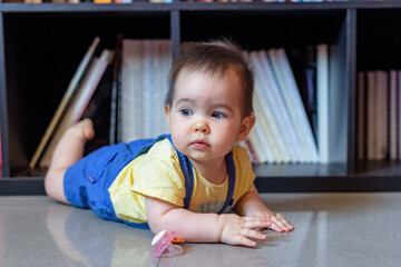 bebe niña pequeña tumbada en suelo delante de biblioteca en hogar - obrazy, fototapety, plakaty