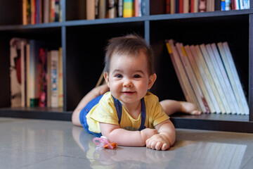 bebe niña pequeña tumbada en suelo delante de biblioteca en hogar - obrazy, fototapety, plakaty