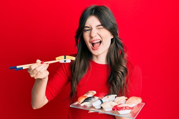 Young beautiful caucasian girl eating sushi using chopsticks winking looking at the camera with sexy expression, cheerful and happy face.