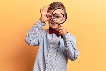 Cute blond kid wearing nerd bow tie and glasses holding magnifying glass smiling happy doing ok sign with hand on eye looking through fingers