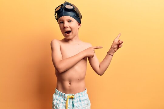 Cute blond kid wearing swimwear and swimmer glasses smiling and looking at the camera pointing with two hands and fingers to the side.