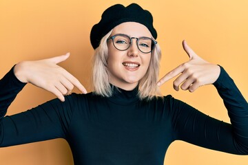 Young blonde girl wearing french look with beret looking confident with smile on face, pointing...