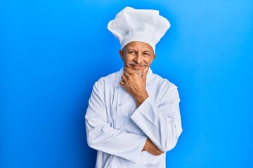 Middle age grey-haired man wearing professional cook uniform and hat looking confident at the camera smiling with crossed arms and hand raised on chin. thinking positive.